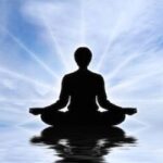 Silhouette of a person meditating in a lotus position on calm water with a bright blue sky and reflection above the water surface.