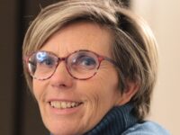 paola-triulzi-foto – Yoga vita e Salute Smiling woman with short gray hair, pink-rimmed glasses, wearing a blue sweater in a portrait shot.