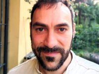 Foto-Ferranti-Roberto – Yoga vita e Salute Close-up of a man with dark hair and a beard outdoors, wearing a light beige shirt.