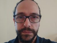 foto-Catellani-Andrea – Yoga vita e Salute Close-up selfie of a man wearing dark glasses, showing his forehead and beard against a plain beige wall.