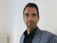 foto-Paolo-Pelloni – Yoga vita e Salute Portrait of a man with dark hair and stubble wearing a blue blazer and black shirt, looking at the camera.