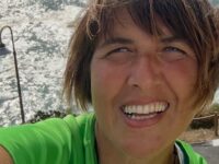 foto-alessandra-begnis-2 – Yoga vita e Salute Close-up selfie of a smiling woman in a bright green shirt, with the sea and rocks in the background.