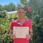 Smiling man in a red polo shirt outdoors, holding a certificate in front of a garden.