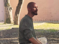IMG-20250914-WA00122-aa – Yoga vita e Salute Side-profile of a bald man with a beard, sitting cross-legged outdoors in a courtyard with trees and a pale wall behind him.